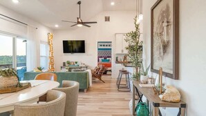 A modern living room with a flat-screen TV, a ceiling fan, a dining area with chairs, a sofa, and a kitchen area with a bar stool.
