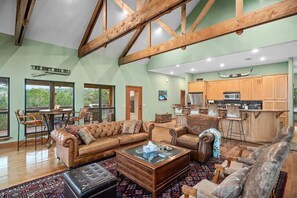 A spacious living room with a large sofa, a coffee table, and a dining area with chairs. There is a kitchen area with wooden cabinets and a bar. - Star House | Private Views • Hill Country Comfort (Wimberley)