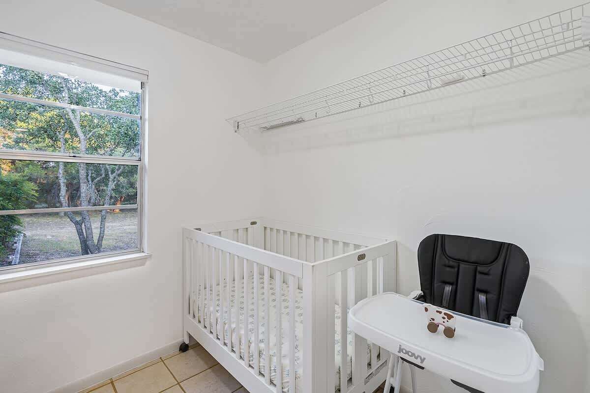 A white crib with a high shelf, a baby seat, and a window with a view of trees.