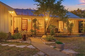 A house with a covered porch, a pathway leading to the entrance, and a small garden with various plants and a tree. - Shady Grove | Hot Tub • Firepit • Pet Friendly (Wimberley)