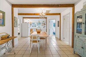 A dining area with a wooden table, white chairs, a chandelier, and a kitchen in the background. - Shady Grove | Hot Tub • Firepit • Pet Friendly (Wimberley)