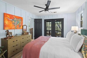 Un dormitorio con cama, ventilador de techo, cómoda, un cuadro y una puerta que da a un espacio exterior.