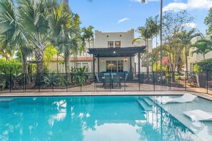 A swimming pool with lounge chairs, a covered patio, and surrounding tropical plants.