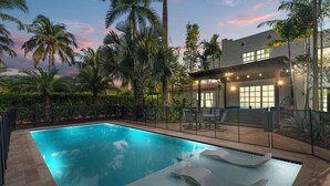 Una piscina con sillas de descanso y una zona de estar cubierta, rodeada de palmeras y una casa con grandes ventanales.