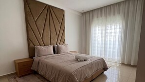 Une chambre à coucher moderne avec un grand lit, une table de chevet, une tête de lit décorative et une grande fenêtre avec des rideaux.