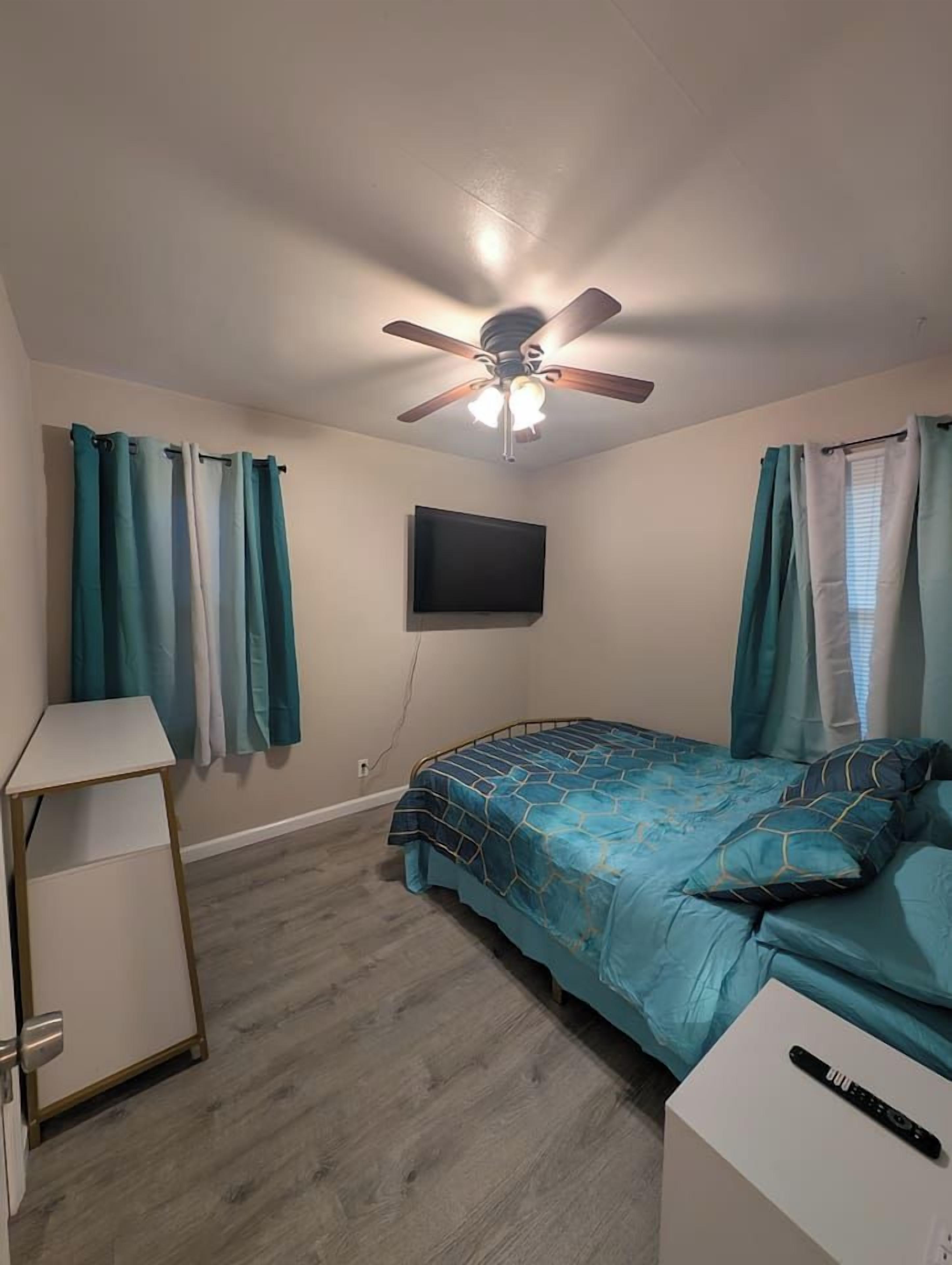 Une chambre à coucher avec un lit, un ventilateur de plafond, un téléviseur fixé au mur et un bureau.