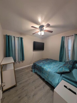 A bedroom with a bed, a ceiling fan, a TV mounted on the wall, and a desk.