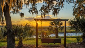 Een houten schommel met een bank eronder, omgeven door bomen en uitzicht op een waterpartij tijdens de zonsondergang.