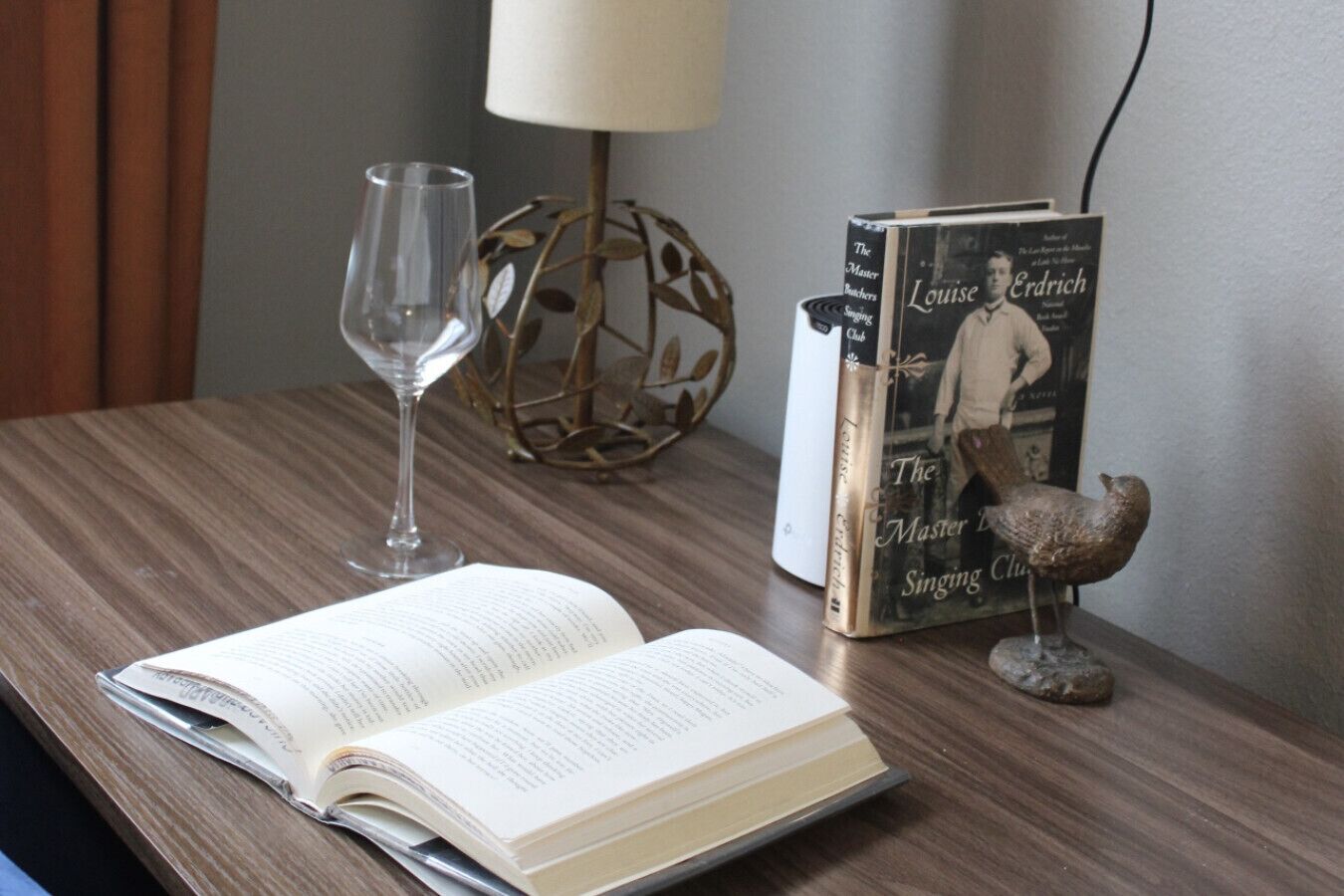 A book titled "The Master Singers" by Louise Erdrich on a wooden table, accompanied by a wine glass and a decorative bird figurine.