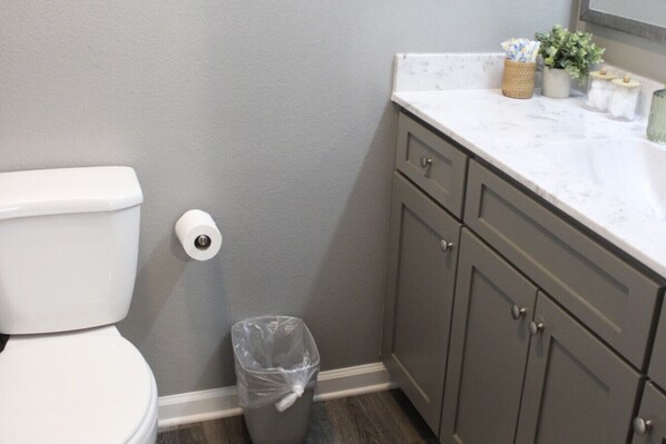 A bathroom with a toilet, a trash can, and a marble countertop with cabinets.