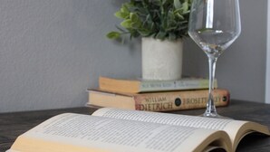 Un libro abierto sobre una superficie de madera, con un vaso de agua transparente a su lado, una planta en maceta y una pila de libros al fondo.