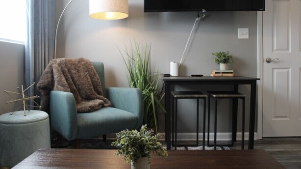 Una sala de estar con un sillón azul, una mesita redonda, una mesa de centro, un televisor instalado en la pared y una lámpara.