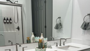 A bathroom with two sinks, a shower, and a large mirror. There are toiletries on the counter and towels hanging on the wall. - •Charming One-Level Home • Feels Like Home (Menifee)