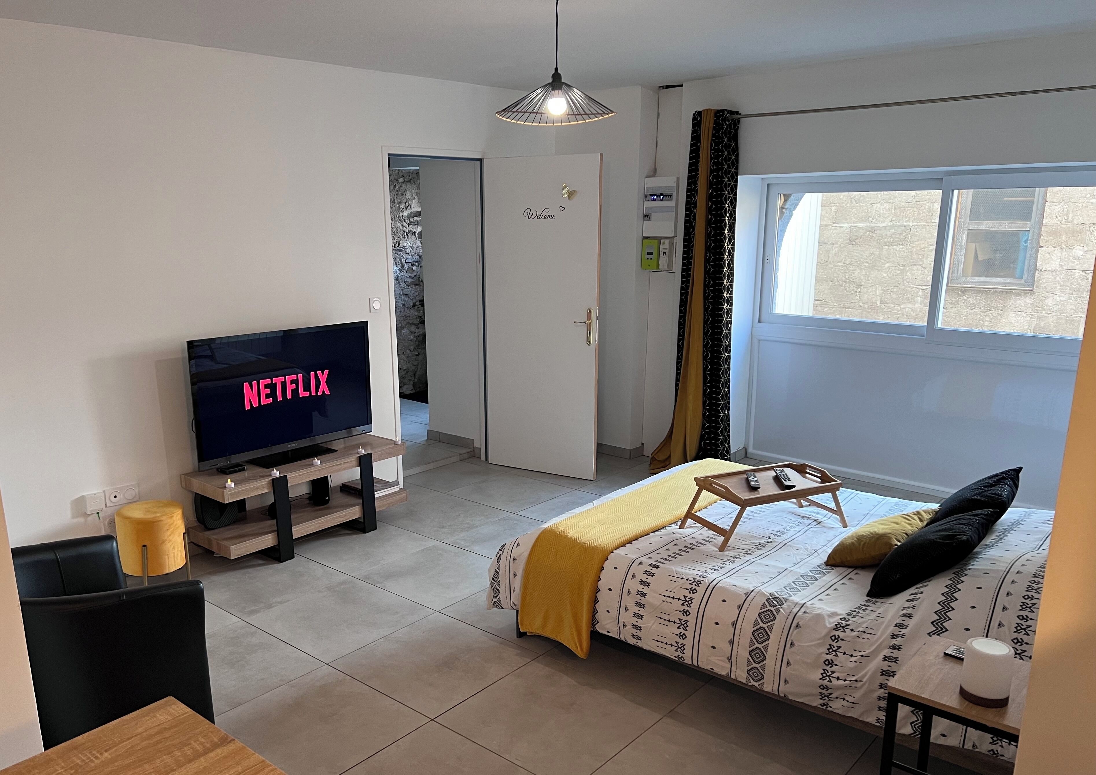 A modern bedroom with a bed, a TV displaying 'NETFLIX', a bedside table, and a window with curtains.