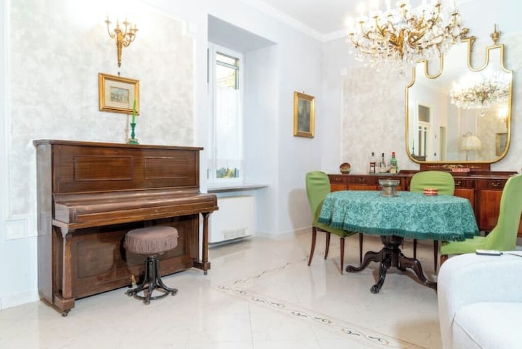A room with a piano, a dining table with chairs, a mirror, and a chandelier.
