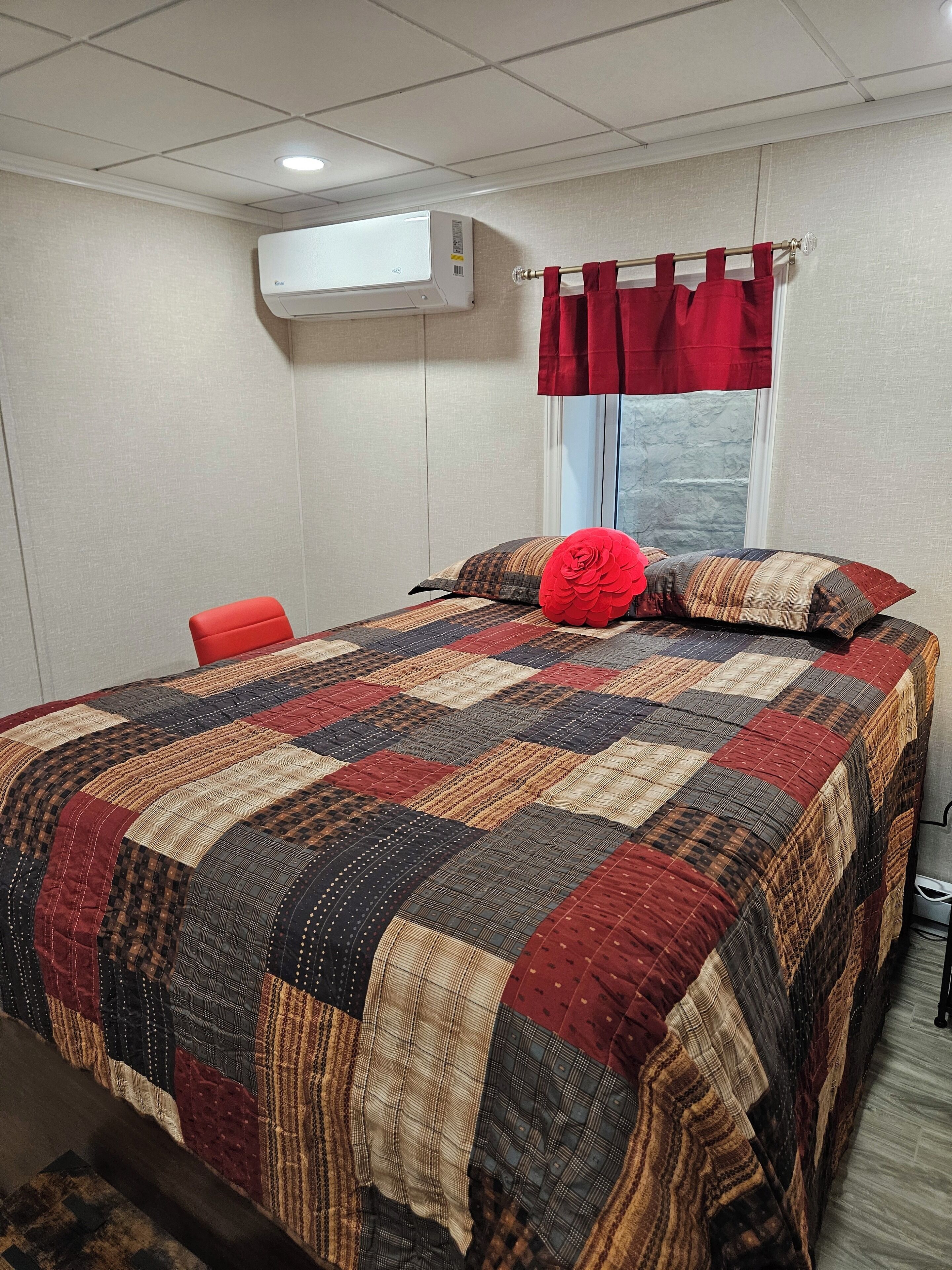 Un dormitorio con una cama grande, una silla roja, una ventana con cortinas rojas y un equipo de aire acondicionado en la pared.