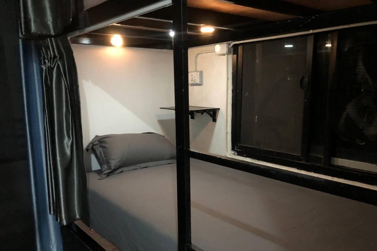 Una cama individual con un almohadón gris en una habitación con ventana, toma de corriente y estante fijado a la pared.