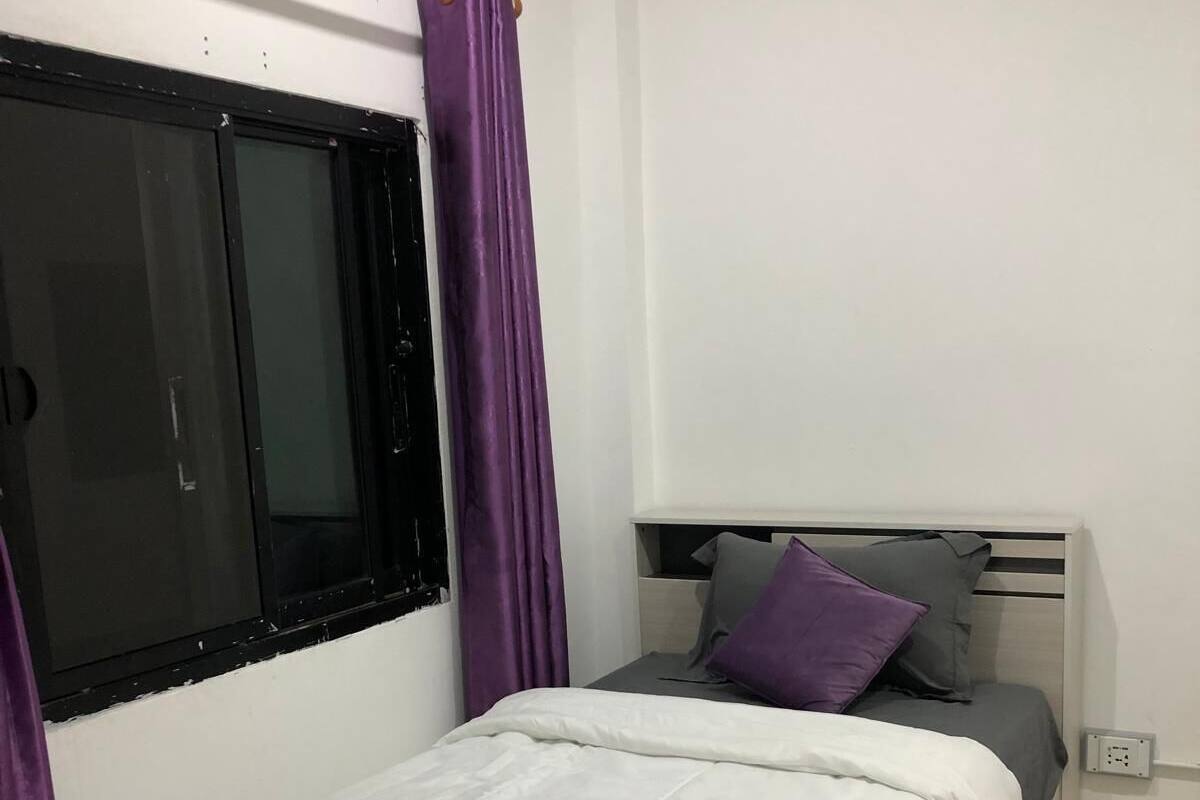 Un dormitorio con cama, una ventana con cortinas moradas, una mesita de noche con lámpara y un enchufe.