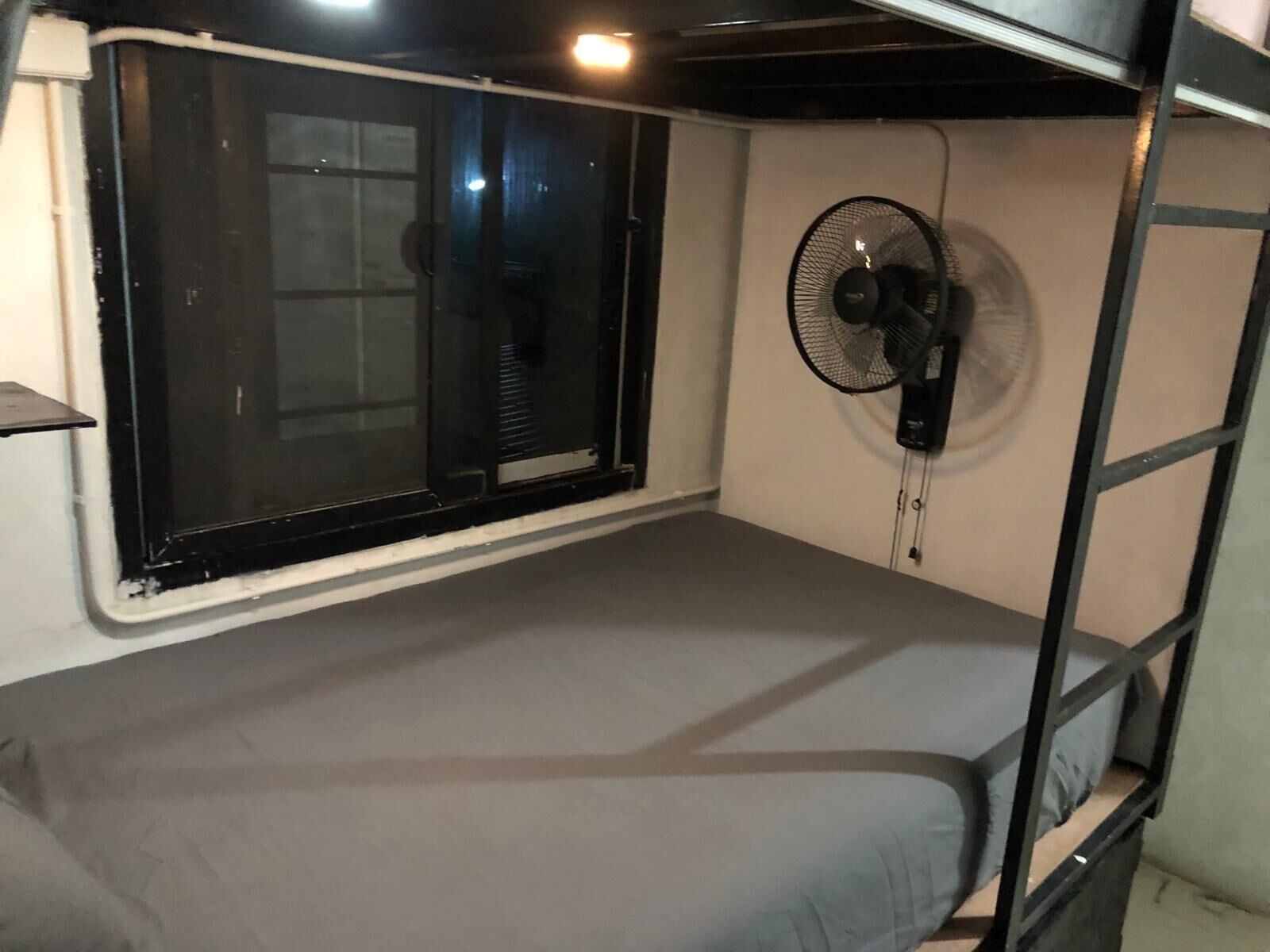 A single-bed room with a window, a wall-mounted fan, and a small shelf.