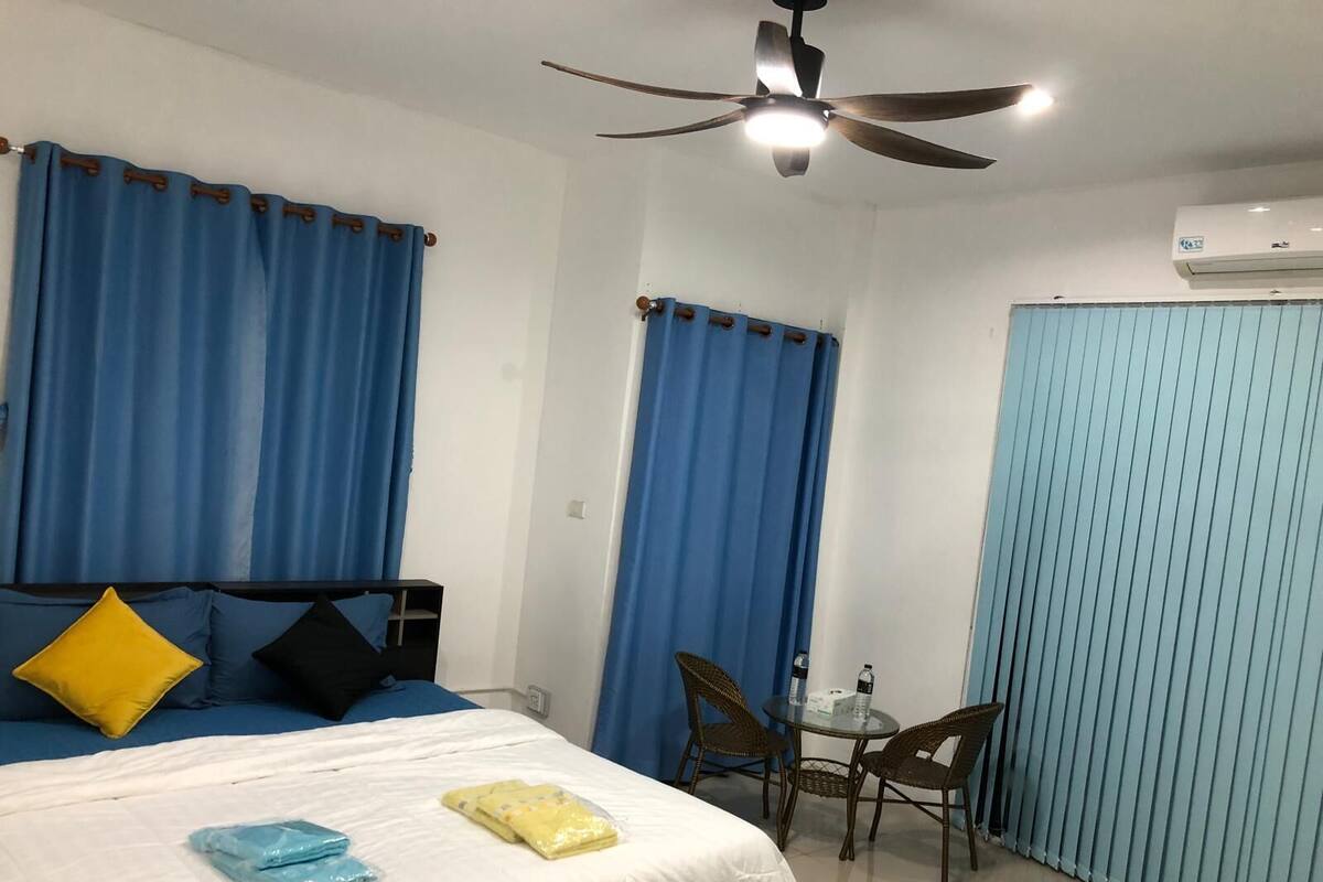 Una habitación de hotel con cama, una mesita con sillas, un ventilador de techo y cortinas azules.