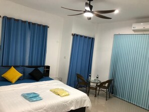 Une chambre d’hôtel avec un lit, une petite table et des chaises, un ventilateur de plafond et des rideaux bleus.