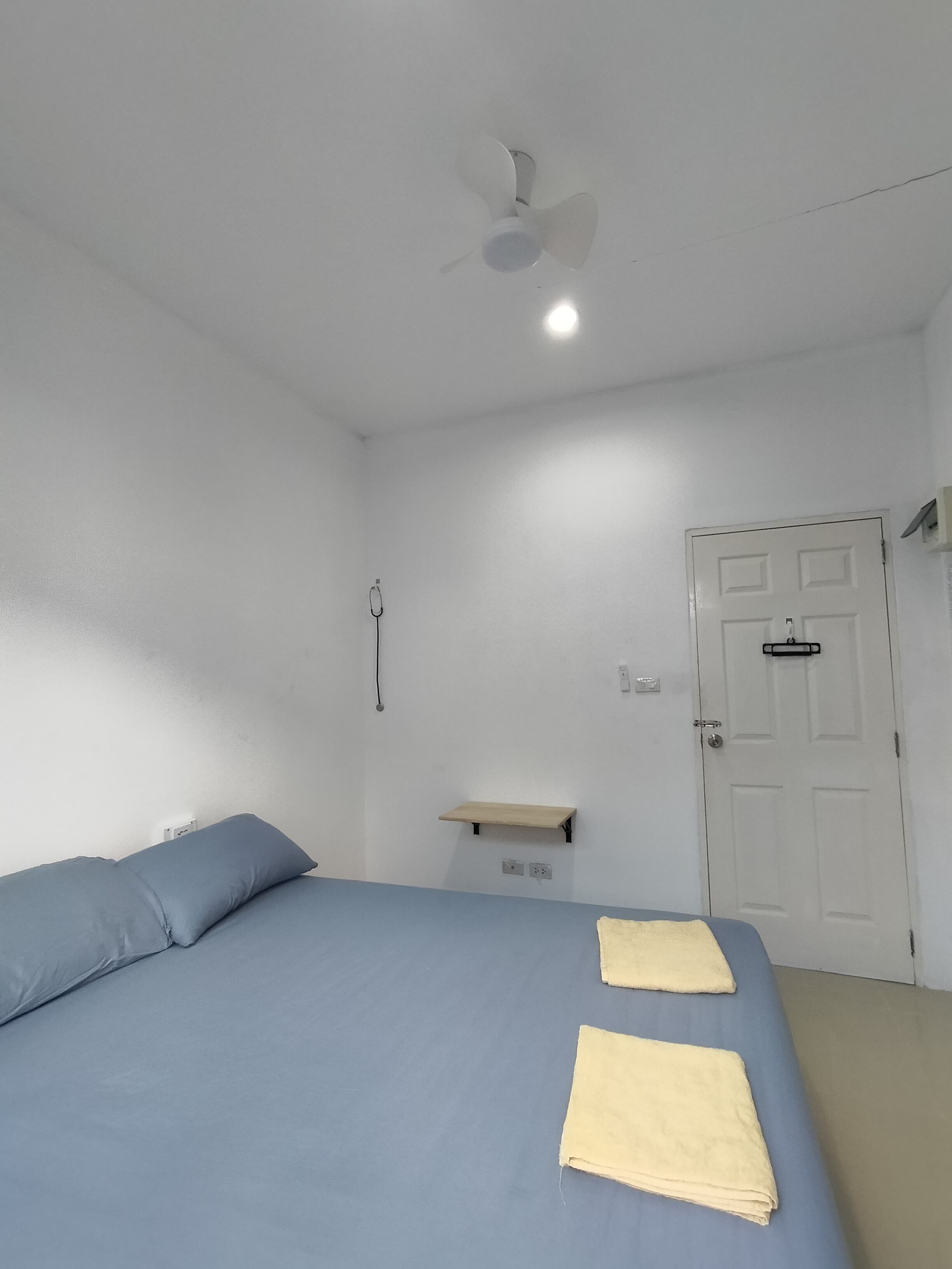 Una habitación con cama, ventilador de techo, una puerta y un estante pequeño.