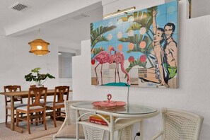 A dining area with a round glass table, white wicker chairs, a hanging light fixture, and a wall mural featuring flamingos and tropical leaves. - ParkView Palms, Mid-Century Home, Sleeps 6, Pets (Mount Dora)
