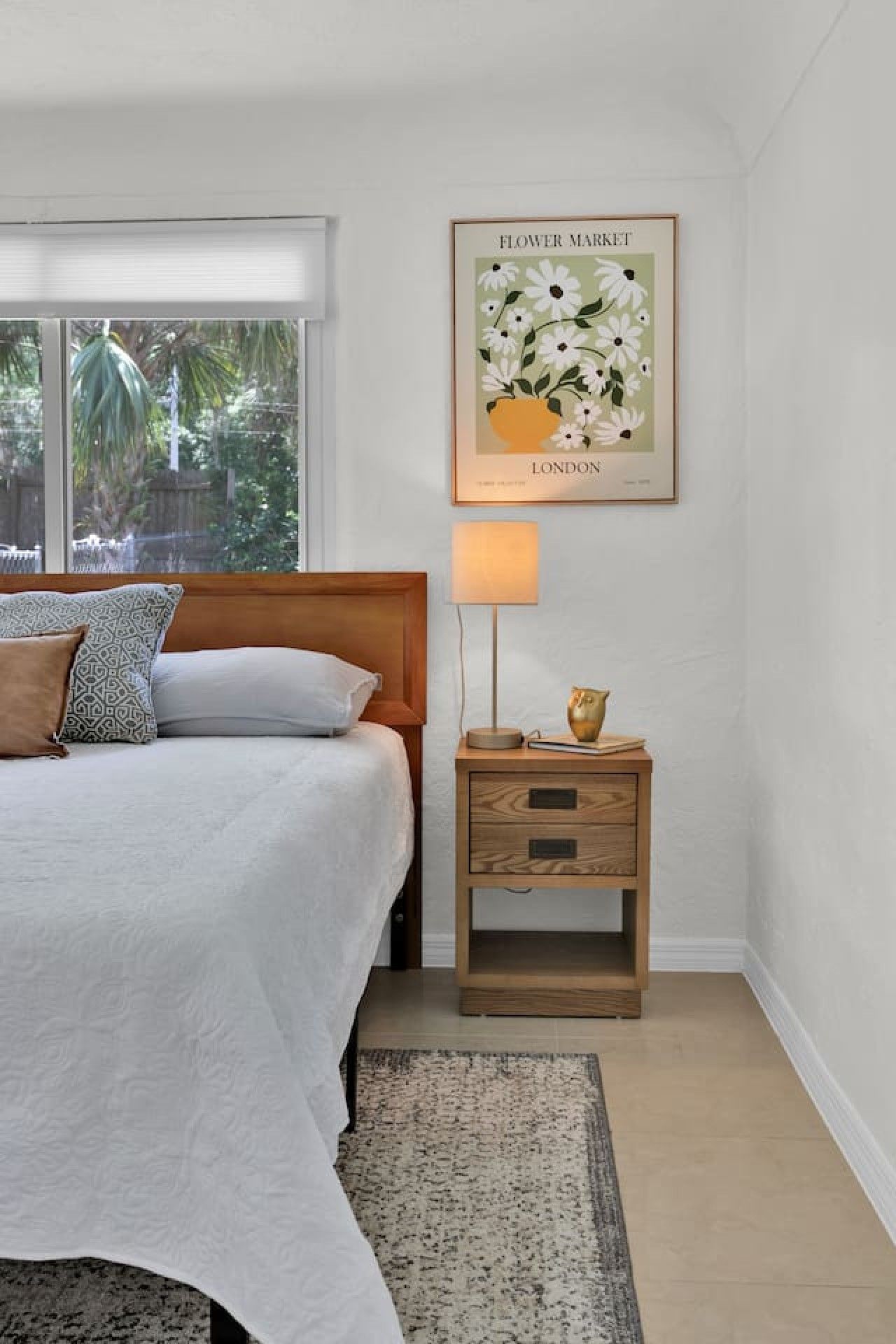 A bedroom with a bed, a nightstand, a lamp, a framed picture, and a window with a view of greenery.