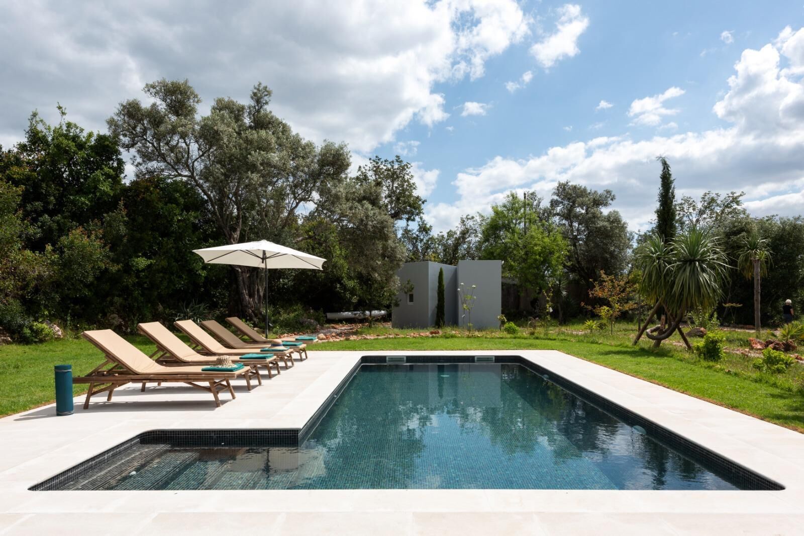 A swimming pool with lounge chairs and umbrellas, surrounded by greenery and a clear sky.