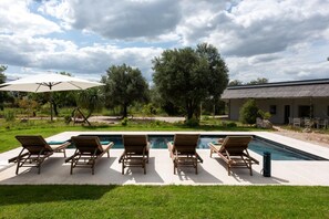 En pool med liggestole, parasol og en bygning i baggrunden.
