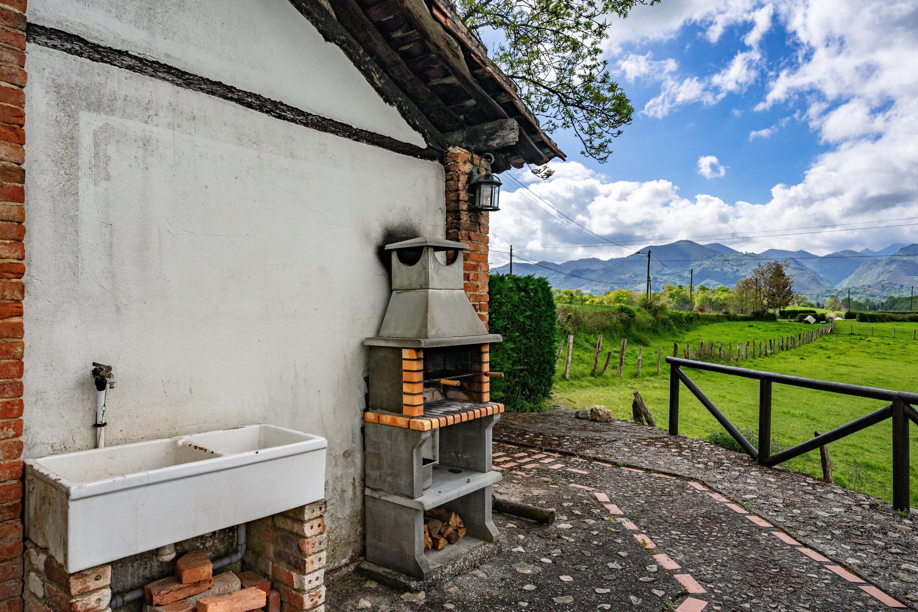 Una cocina rústica al aire libre con horno de ladrillo, un lavamanos y un sendero de piedra que lleva a una zona cercada con vistas a las montañas.