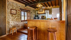 A rustic kitchen with stone walls, wooden beams, a bar area with stools, and a stone fireplace.