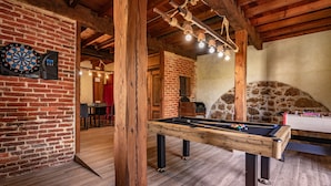 A billiards table in a rustic room with exposed wooden beams, a dartboard, and a stone wall.