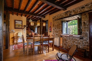 Un interior rústic amb una paret de pedra, bigues de fusta al sostre, una zona de menjador amb cadires i taula, i una finestra amb vista a vegetació.
