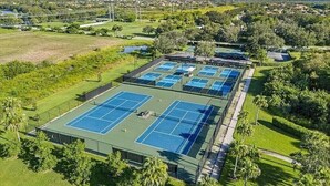 An aerial view of a tennis facility with multiple courts, surrounded by landscaped areas and trees.