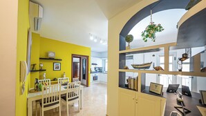 A bright kitchen with yellow walls, a dining area with a white table and chairs, and a workspace with shelves and a hanging plant. - El Charco Azul (San Andrés y Sauces)