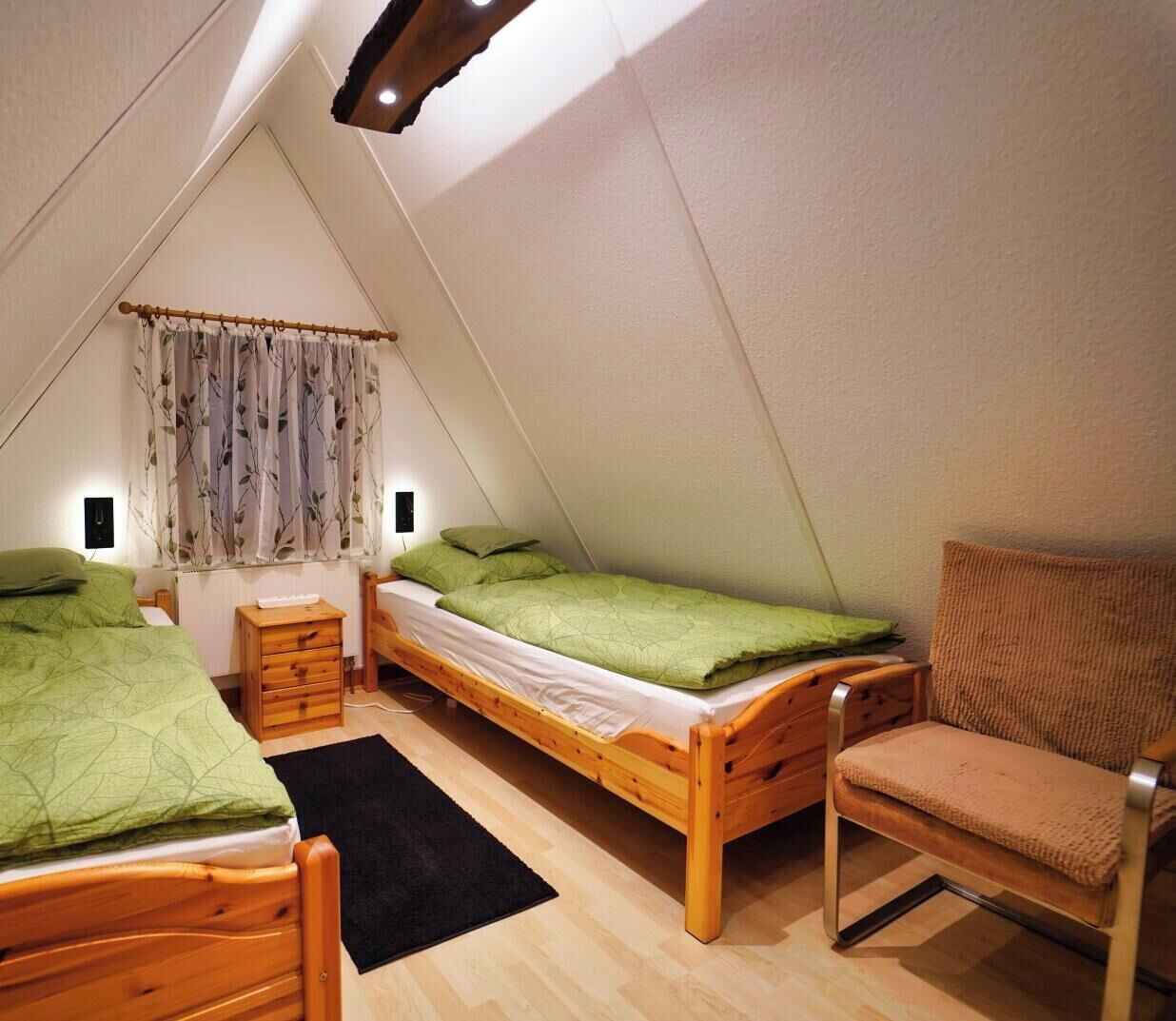 Une petite chambre mansardée de forme triangulaire, avec deux lits en bois, une chaise et une petite table.
