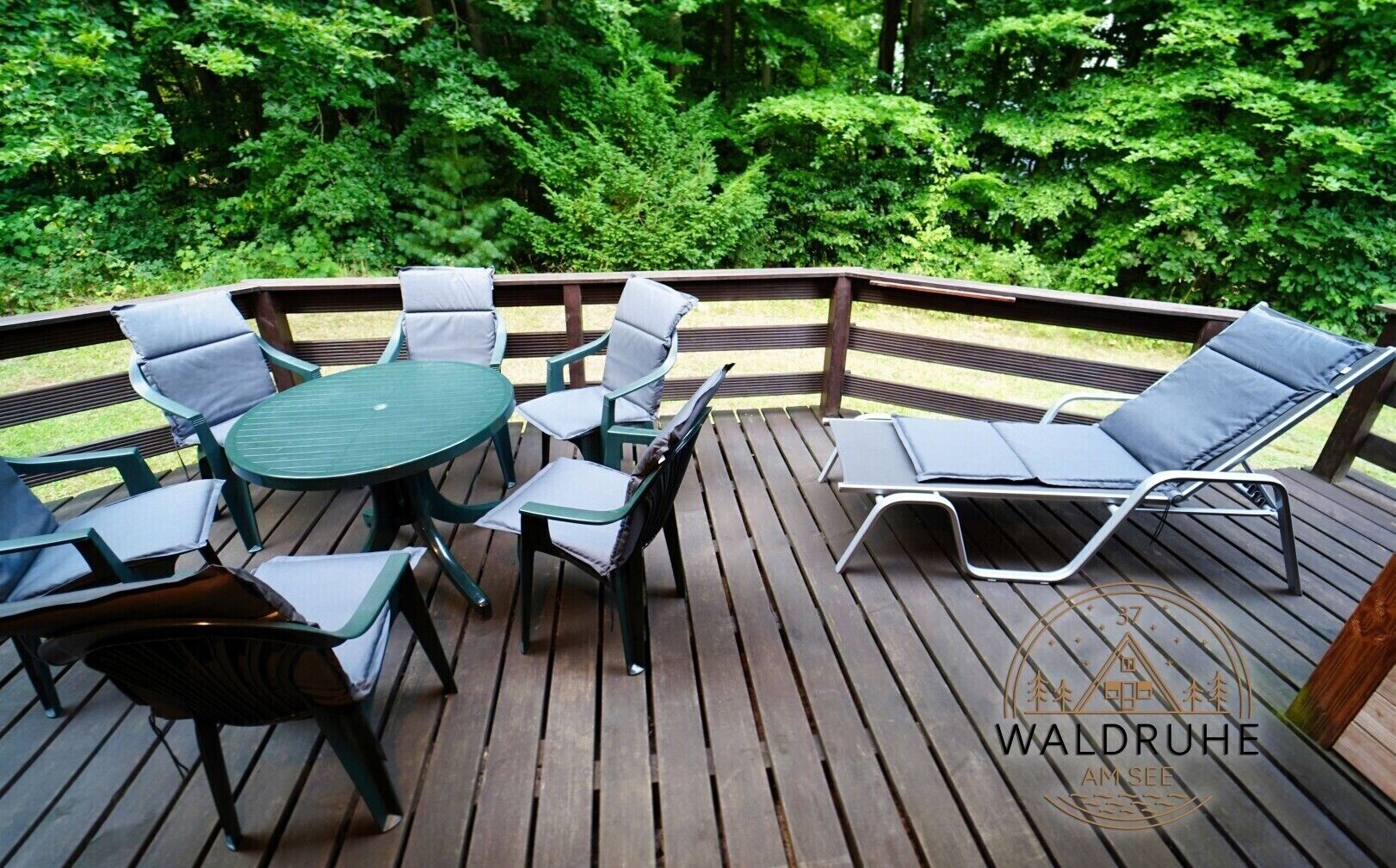 A wooden deck with a round table and chairs, a lounge chair, and a wooden railing.