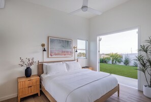 Un dormitorio con cama, mesitas de noche, ventilador de techo, ventana y vista a un jardín verde.