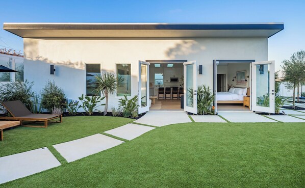 Una casa moderna con un amplio patio trasero, un comedor y un dormitorio visibles a través de puertas abiertas.