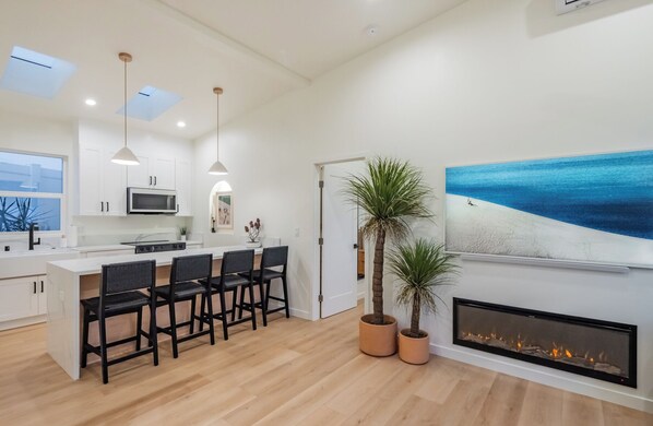 A modern kitchen with white cabinets, a central island with bar stools, a microwave, and pendant lighting. There's a fireplace with a fire, a large beach-themed painting, and two potted plants.