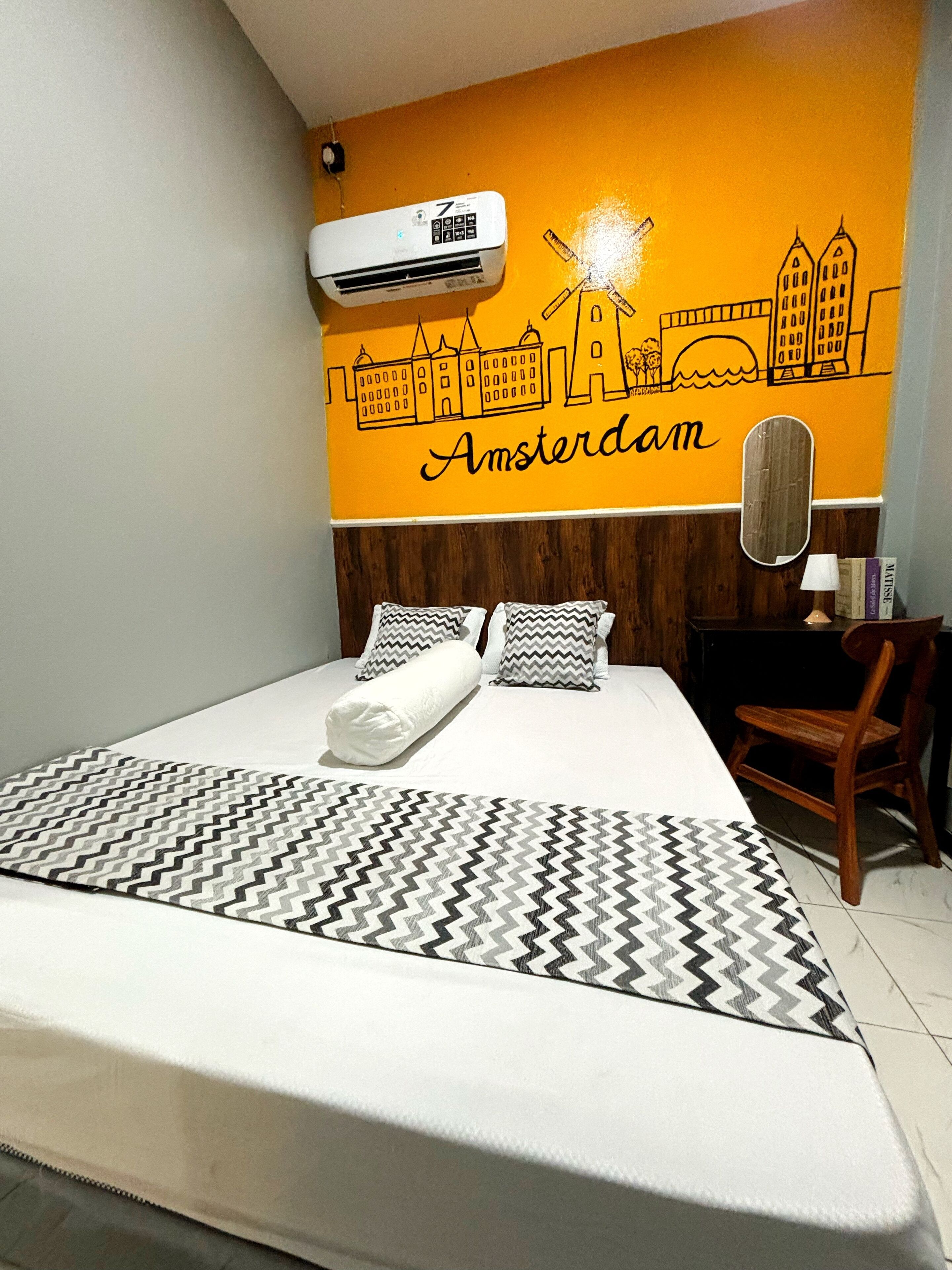 A hotel room with a bed, a wall mural of Amsterdam, a wooden headboard, a mirror, and a desk with a chair.