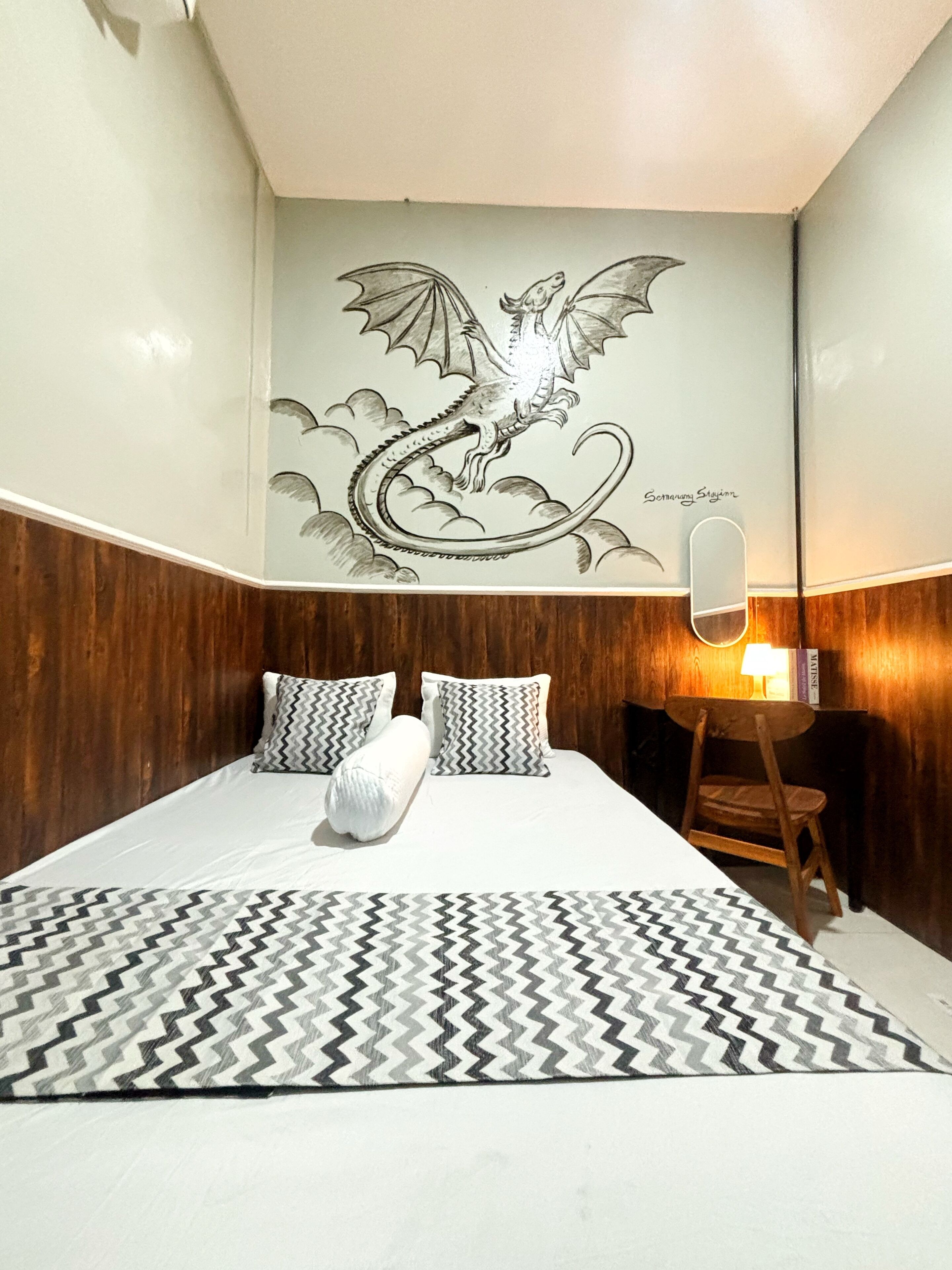 A hotel room with a bed, a patterned rug, a wooden headboard, a wall-mounted mirror, and a dragon mural.