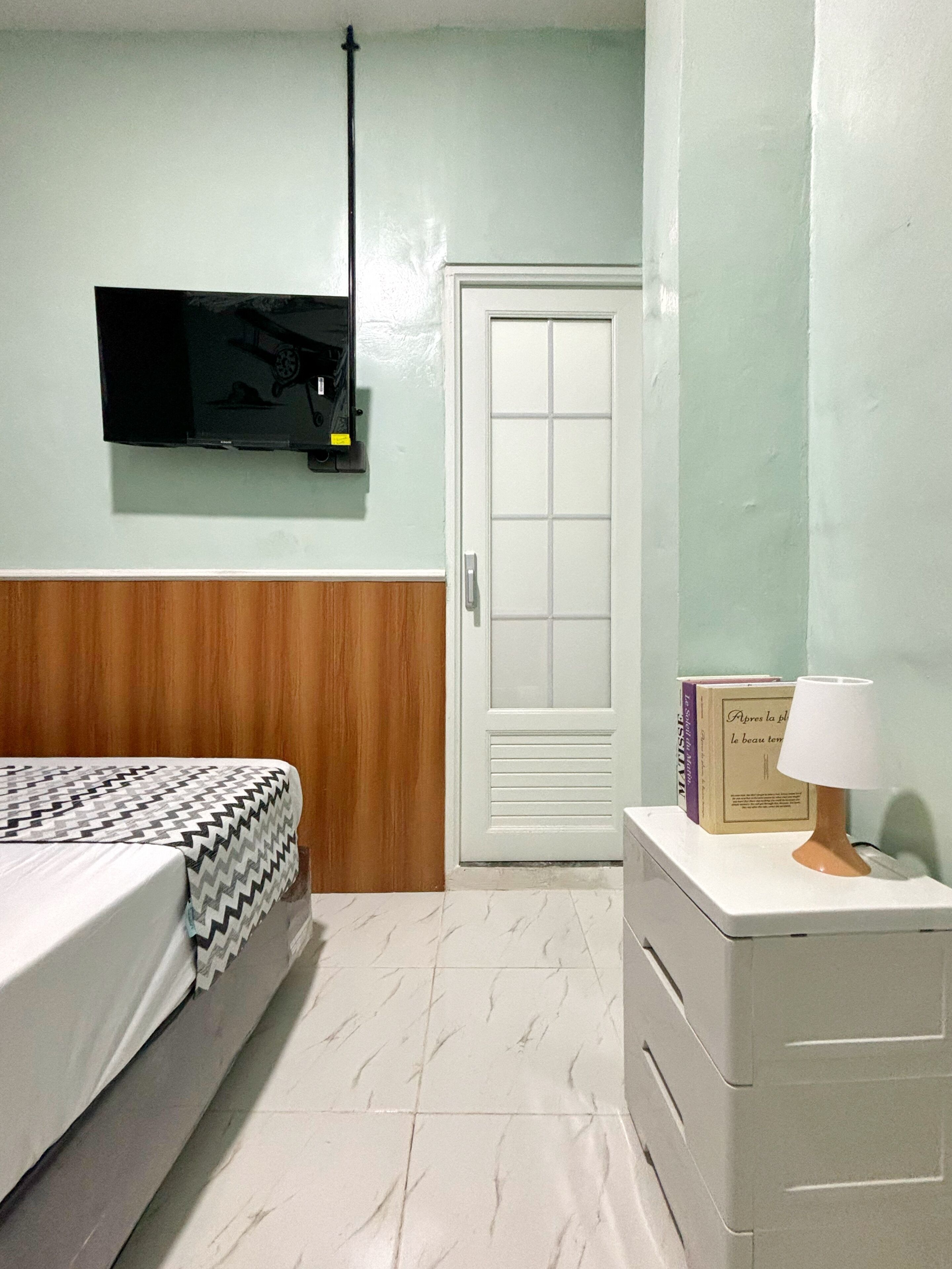 A bedroom with a bed, a nightstand, a lamp, a television mounted on the wall, and a door.
