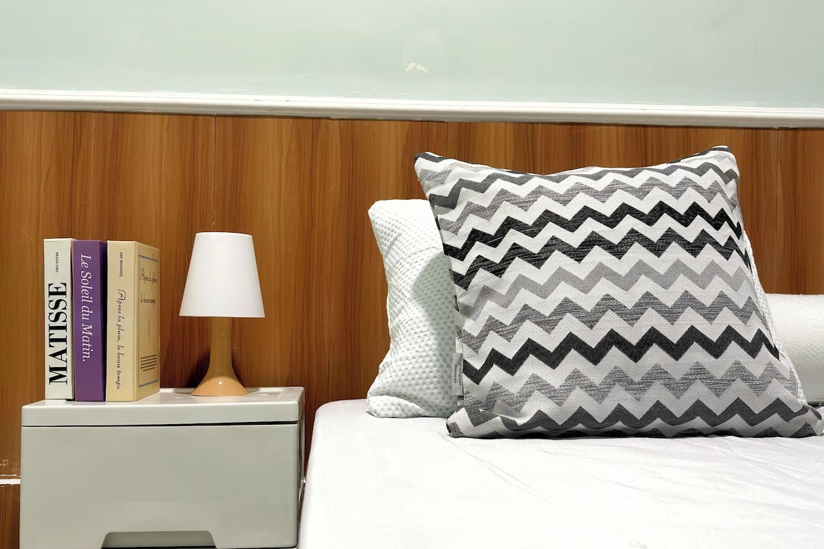 A bedroom with a bed, a nightstand with books, a lamp, and a wall with a name.