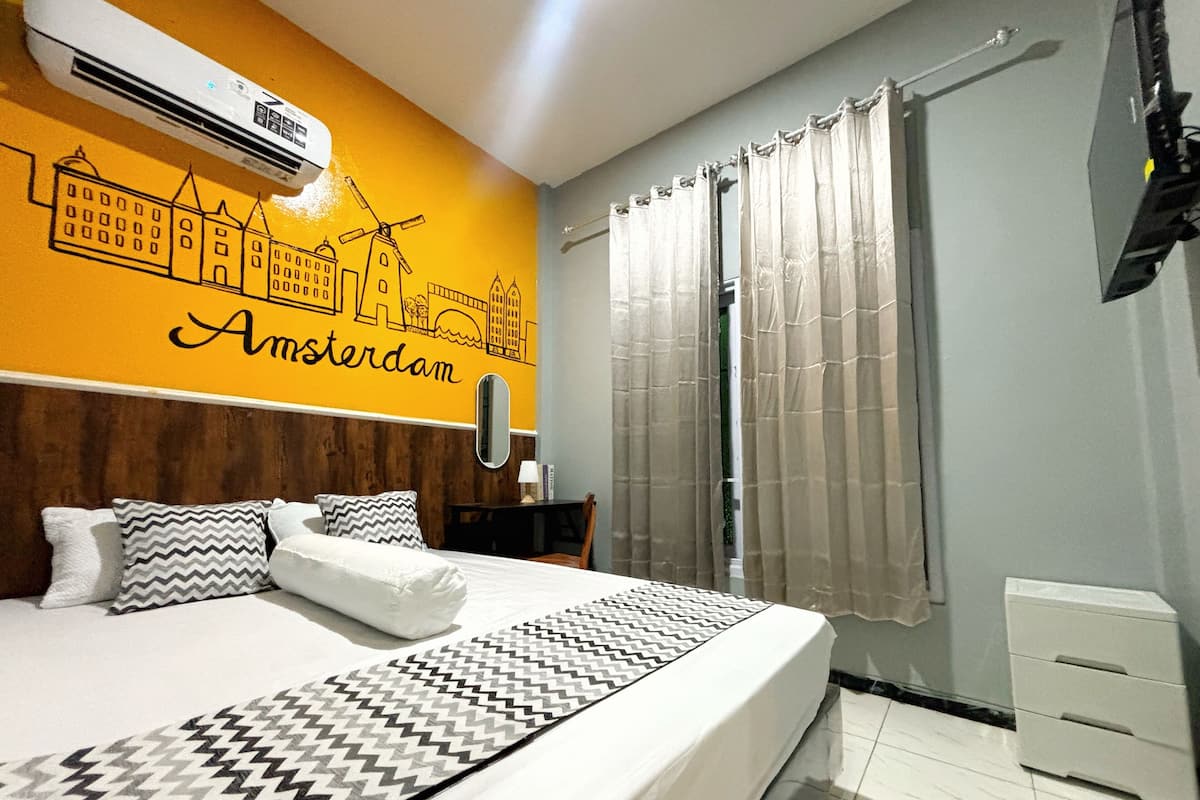 A hotel room with a bed, a wooden headboard, a wall mural of Amsterdam, a window with curtains, and a white bedside table with a lamp.