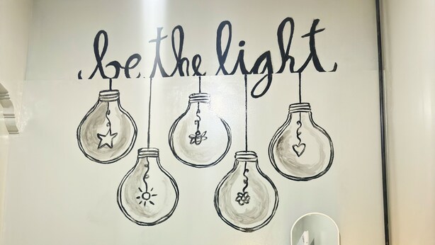 A bedroom with a bed, two pillows, a bedside table with a lamp, and a wall with light bulbs and the phrase "be the light." - Semarang Co Stay (Semarang)