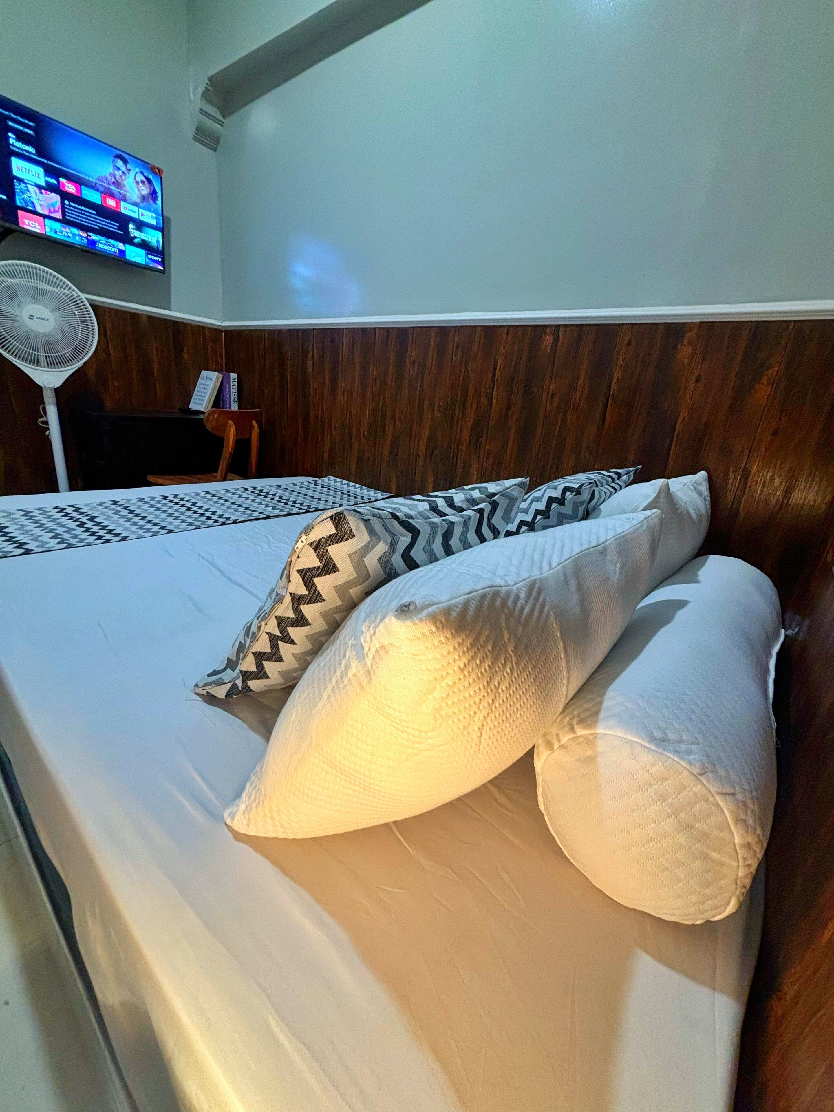 Una cama bien hecha con almohadillas estampadas y un cabecero de madera. Un televisor de pantalla plana está montado en la pared sobre la cama.