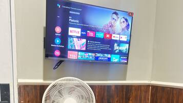 A room with a television mounted on the wall displaying various streaming services, a white standing fan, a wooden chair, and a small table with books.