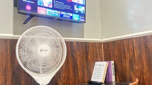 A room with a flat-screen TV mounted on the wall displaying various streaming services, a white standing fan, a wooden chair, and a small table with books. - Semarang Co Stay (Semarang)